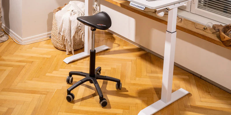 Ergonomic saddle chair on wheels next to a standing desk in a cozy room with wooden flooring.