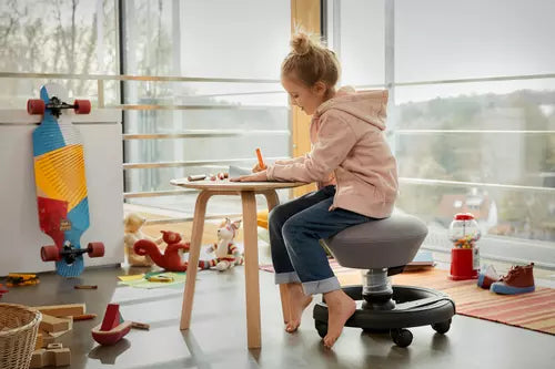 Child sitting on grey Aeris Swoppster active chair at a wooden table, with toys and skateboard in modern children's playroom.